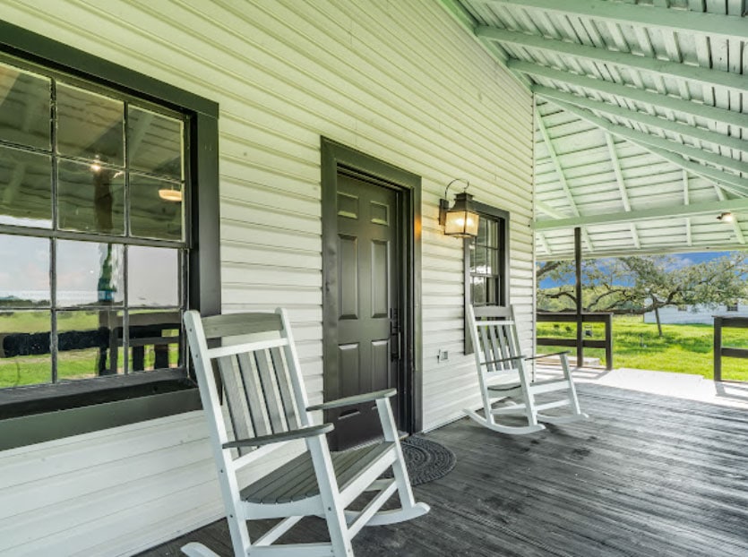 Front porch of a house with two white rocking chairs and a black door in a rural setting.