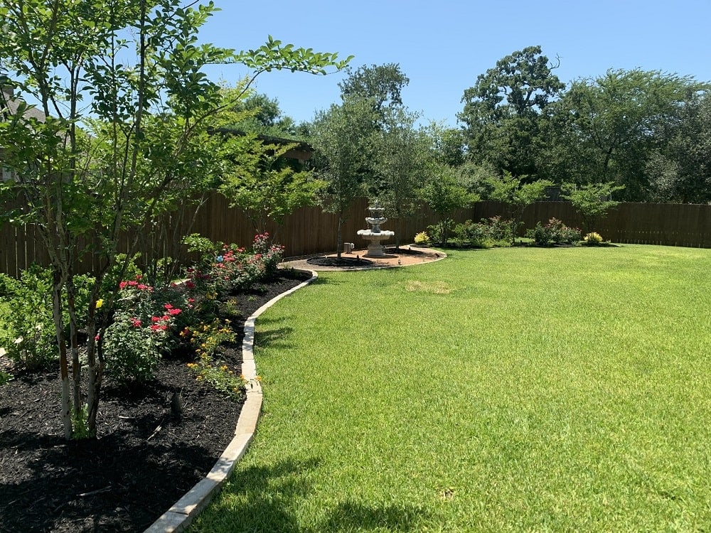 Lush green backyard with flower beds, trees, and a fountain, enclosed by a wooden fence.